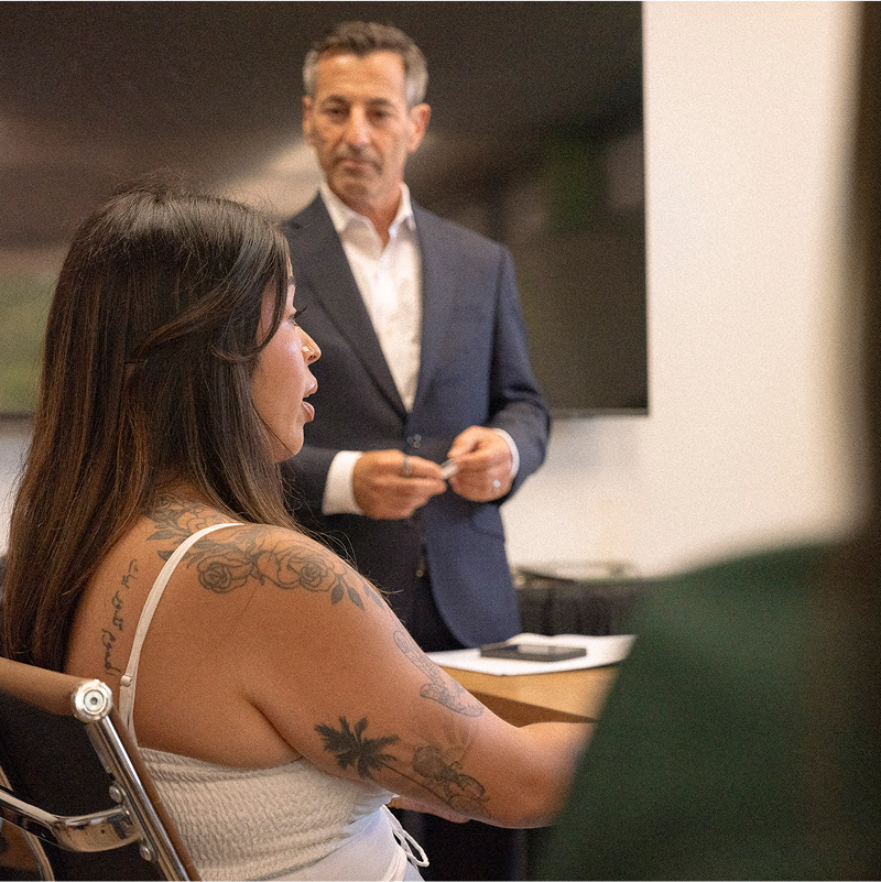 A woman speaks while sitting at a table. A man in a suit stands in the background, listening attentively.