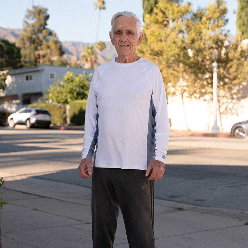 Elderly man in a white long-sleeve shirt and dark pants stands on a sunny suburban street, with trees and houses in the background.