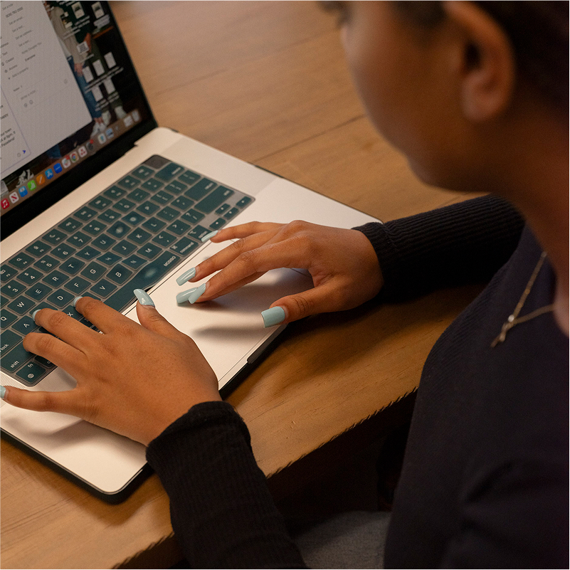 Person typing on a laptop with a wooden table background, wearing a dark sweater and light blue nail polish.