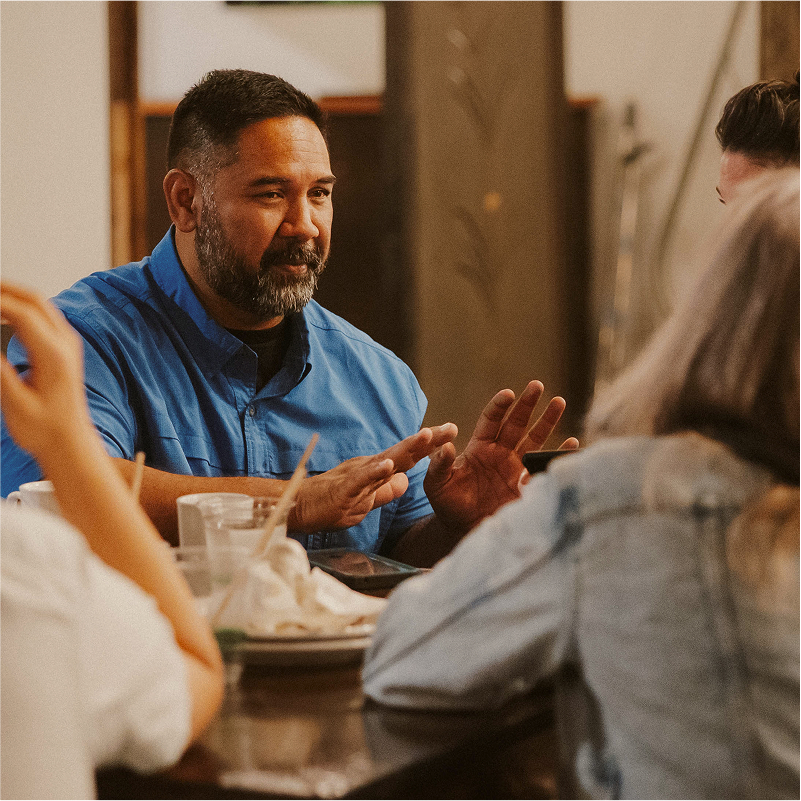Man in a blue shirt talking and gesturing at a table with others, with plates and cups visible in a casual setting.