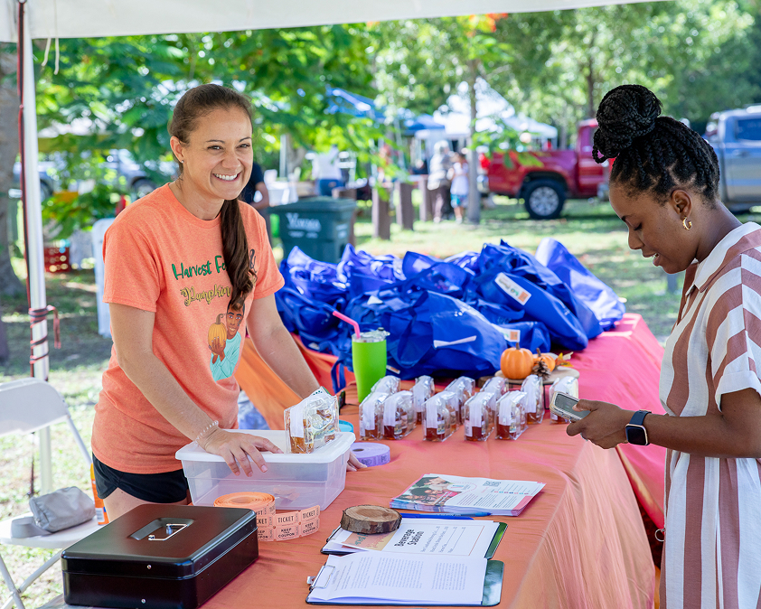 A woman at a booth with orange tablecloth and blue bags smiles at another person. Various items and pamphlets are displayed on the table.
