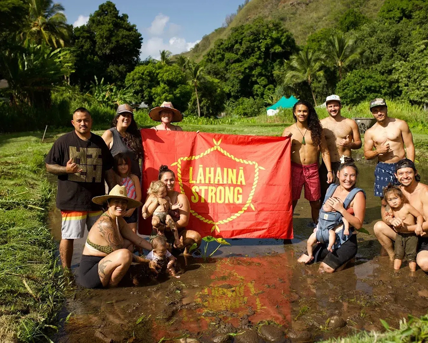 Group of people in a lush, tropical setting holding a red "Lahaina Strong" banner, posing in shallow water.