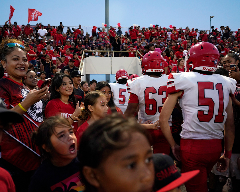 Football players in red uniforms walk through a cheering crowd in a stadium. Fans wear red and black, holding flags and banners.