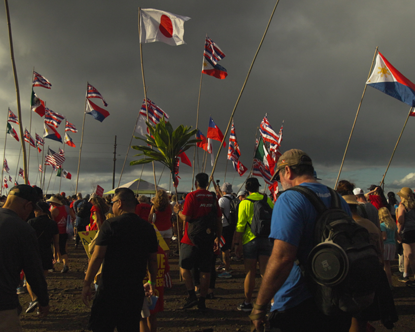 Crowd gathered under a cloudy sky, holding various flags on poles, including Japan, Philippines, and U.S. flags.
