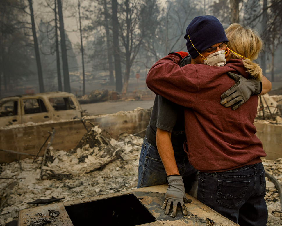 Two people in protective masks and gloves embrace amidst the charred remains of a forest and vehicles, with a somber, smoky backdrop.