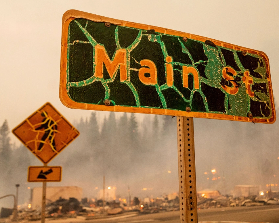 A damaged "Main St" sign stands in front of a hazy, smoke-filled background.
