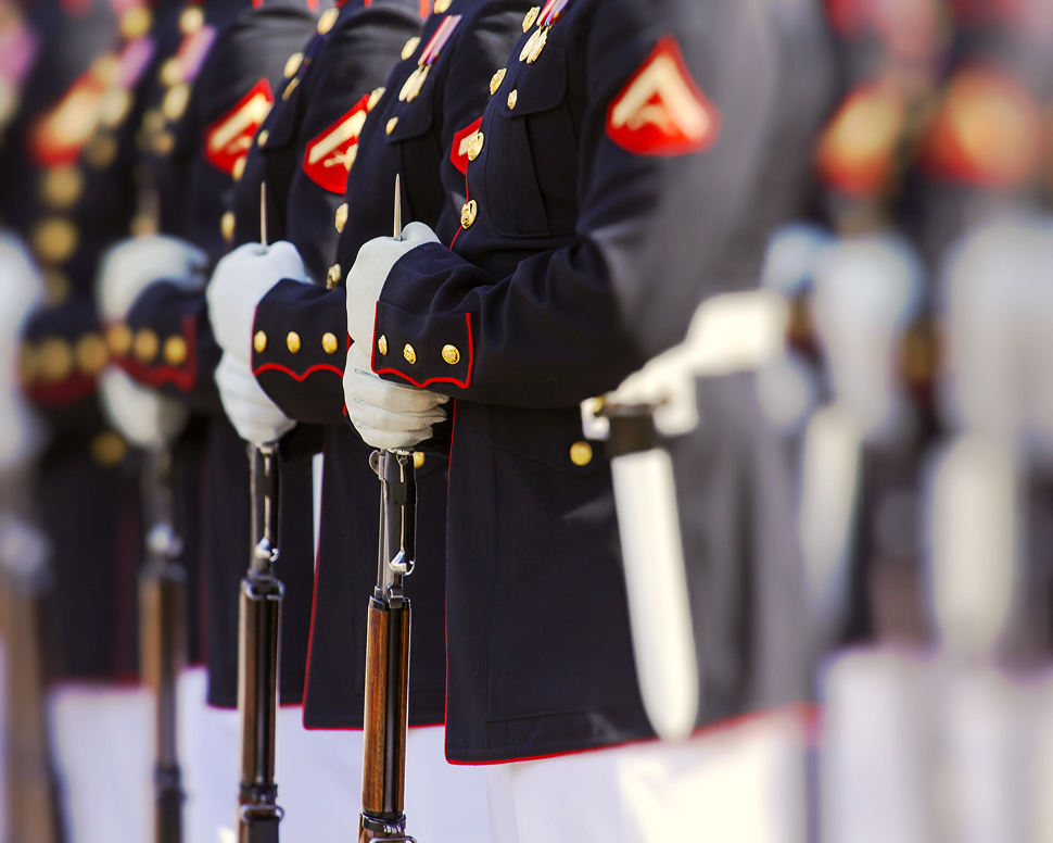 Uniformed soldiers in formation, wearing dark blue jackets with red trim and white gloves, holding rifles vertically in front of them.