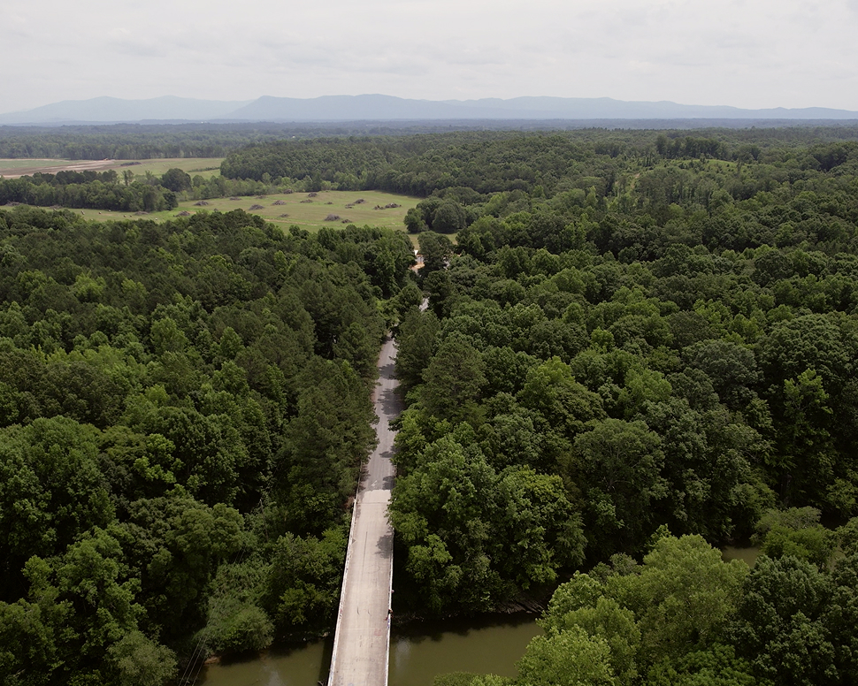 Aerial view of a road cutting through dense green forest, with distant mountains under a cloudy sky.