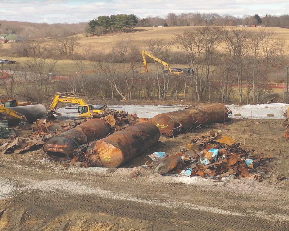 Construction site with rusted, dismantled fuel tanks, excavators, and debris on a dirt landscape. Bare trees and rolling hills in the background.