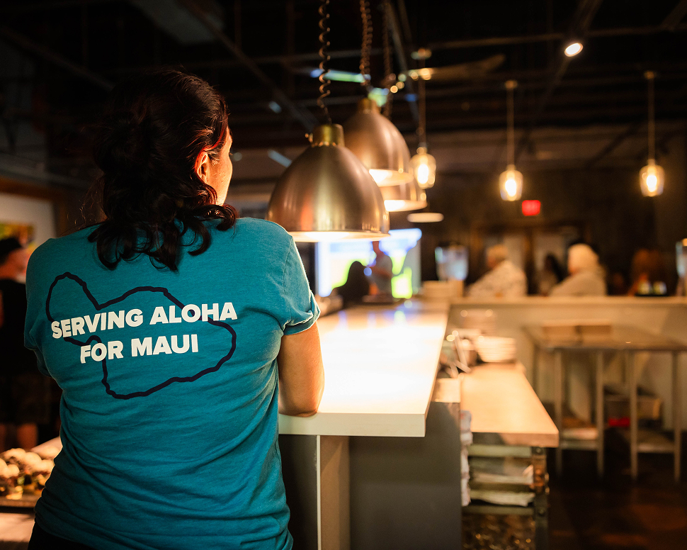 Person in a blue shirt with "Serving Aloha for Maui" printed on the back stands at a counter in a warmly lit restaurant.