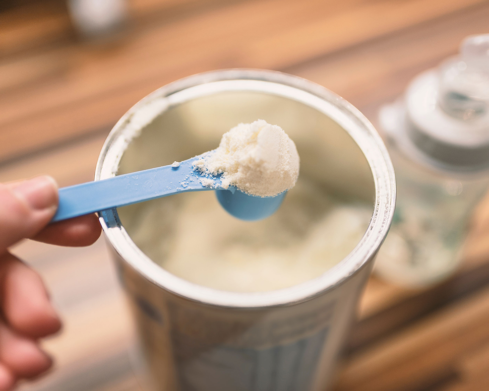 A hand holds a blue scoop of powdered formula above an open container on a wooden surface.