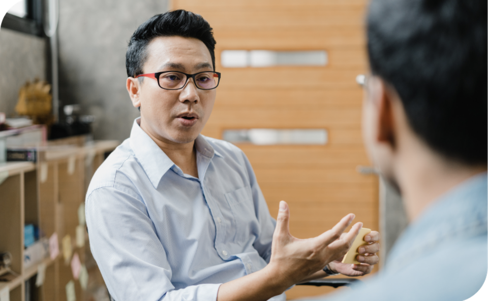A man wearing glasses and a light blue shirt is gesturing while talking to another person in an office setting.