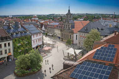 Der Marktplatz von Bayreuth von einem Dach aus gesehen im Sommer – auf Dächern sind Solarpanele grafisch eingefügt, ein Haus hat eine Fassadenbegrünung, ein kleine Tiny Forest Insel ist als Begrünung zu sehen und ein weißes Sonnensegel spendet Schatten.
