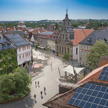 Der Marktplatz von Bayreuth von einem Dach aus gesehen im Sommer – auf Dächern sind Solarpanele grafisch eingefügt, ein Haus hat eine Fassadenbegrünung, ein kleine Tiny Forest Insel ist als Begrünung zu sehen und ein weißes Sonnensegel spendet Schatten.
