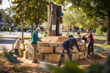 Menschen arbeiten an einer von großen Steinen umfassten kleinen Insel die ein Insektenbiotop werden soll. Im Hintergrund eine Straße.