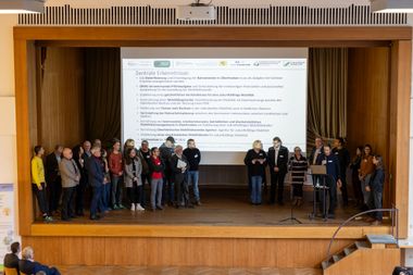 Menschen stehen auf einer Bühne, auf der Leinwand ist eine Folie mit der Überschrift "zentrale Erkenntnisse" zu sehen.