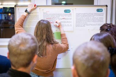 Eine Frau schreibt Anmerkungen mit Rotsstift auf ein ausgedrucktes Blatt mit Text, das an einer Stellwand angeheftet ist. Im Vordergrund sind mehr Menschen zu sehen, die zusehen.