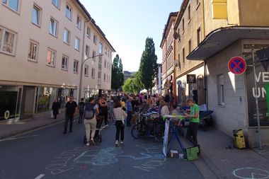 People gather on a city street with chalk drawings on the pavement and stalls lining the sides under a clear blue sky.