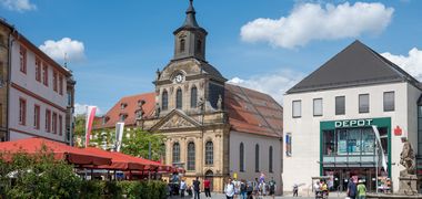 Blick auf einen Platz in der Innenstadt Bayreuths, großes altes Gebäude mit Uhr in der Mitte, links Sonnenschirme und grüne Pflanzkübel eines Gastrobetriebes.