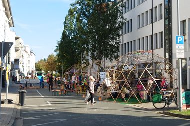 People gather on a city street with chalk drawings on the pavement and stalls lining the sides under a clear blue sky.