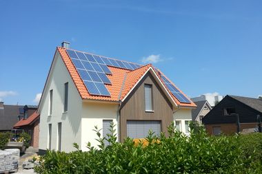 Solar Paneele auf dem roten Ziegeldach eines Einfamilienhauses, das Haus ist eingerahmt von einer grünen Hecke und blauem Himmel.