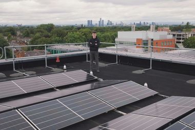 Ein junger Mann steht auf einem Flachdach vor Solarpanelen, im Hintergrund Bürogebäude, grüne Bäume und ein paar Hochhäuser.