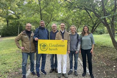 Gruppenfoto von sechs Menschen die ein gelbes Banner "Klimastimmen Bayreuth" halten in einem grünen Park.