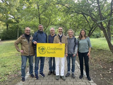 Gruppenfoto von sechs Menschen die ein gelbes Banner "Klimastimmen Bayreuth" halten in einem grünen Park.