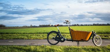Ein Lastenrad auf einem Radweg, im Hintergrund idyllische grüne Felder, ein Kirchturm und blauer Himmel mit ein paar Wolken.