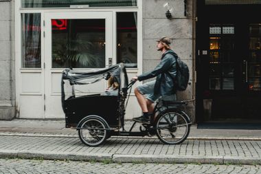 Ein junger Mann fährt ein Lastenrad mit Kind im Korb auf einem gepflasterten Radweg in der Stadt.
