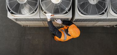 Sicht von oben auf einen Arbeiter in orangener Warnweste und Helm, der einen von mehreren Ventilatoren einer Lüftung auf einem Dach inspiziert.