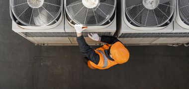 Sicht von oben auf einen Arbeiter in orangener Warnweste und Helm, der einen von mehreren Ventilatoren einer Lüftung auf einem Dach inspiziert.