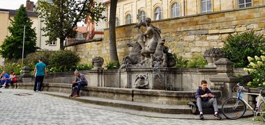 Menschen sitzen an einem Springbrunnen an der Opernstraße in Bayreuth.