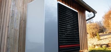 Das Außengerät einer Wärmepumpe steht vor einem Haus mit Holzfassade.