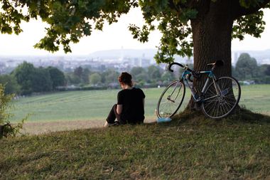 Eine Frau sitzt unter einem Baum, an dem ein Fahrrad lehnt. Im Hintergrund ein weites Feld und eine Stadt.
