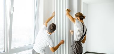 Zwei Handwerker installieren einen hohen Heizkörper in einer lichtdurchfluteten Wohnung.