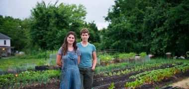Zwei Menschen stehen lächelnd in Gummistiefeln und Latzhose in ihrem Gemüsegarten. Mehrer Pflanzenreihen und Bewässerungsschläche sind zu erkennen, die Erde hat einen reichen, dunklen Ton. Im Hintergrund grüne Bäume und ein Haus.
