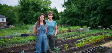 Zwei Menschen stehen lächelnd in Gummistiefeln und Latzhose in ihrem Gemüsegarten. Mehrer Pflanzenreihen und Bewässerungsschläche sind zu erkennen, die Erde hat einen reichen, dunklen Ton. Im Hintergrund grüne Bäume und ein Haus.