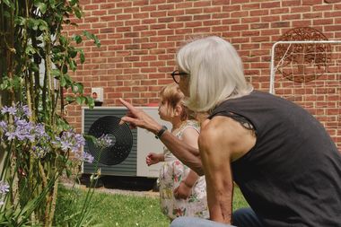 Eine ältere Frau und ein Kleinkind im Garten, die Frau zeigt auf Pflanzen. Im Hintergrund ist eine Wärmepumpe an einer Hauswand zu sehen.