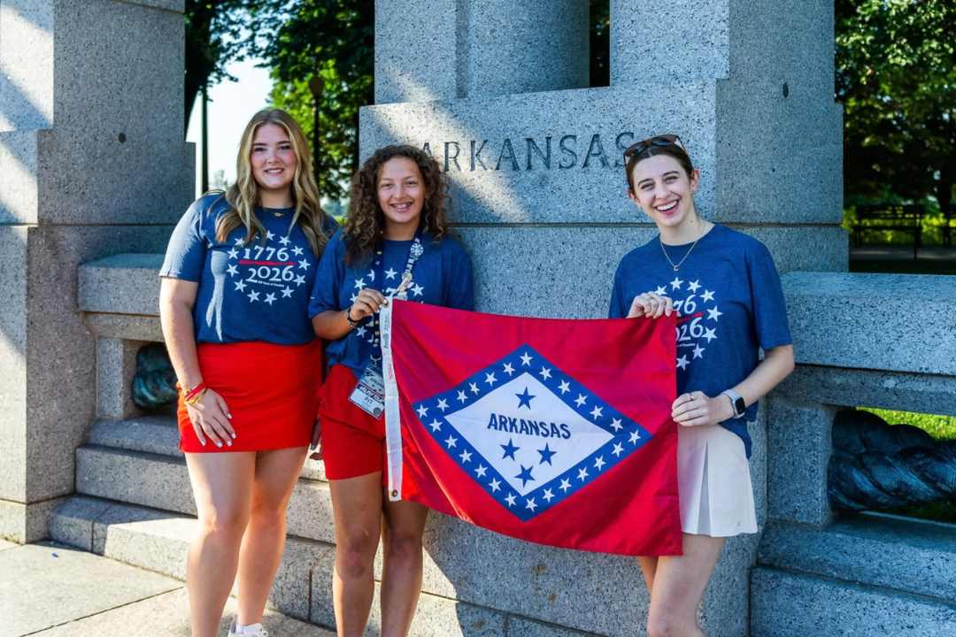 Three people smiling, wearing blue shirts and red skirts, hold an Arkansas flag in front of a stone structure engraved with "ARKANSAS."