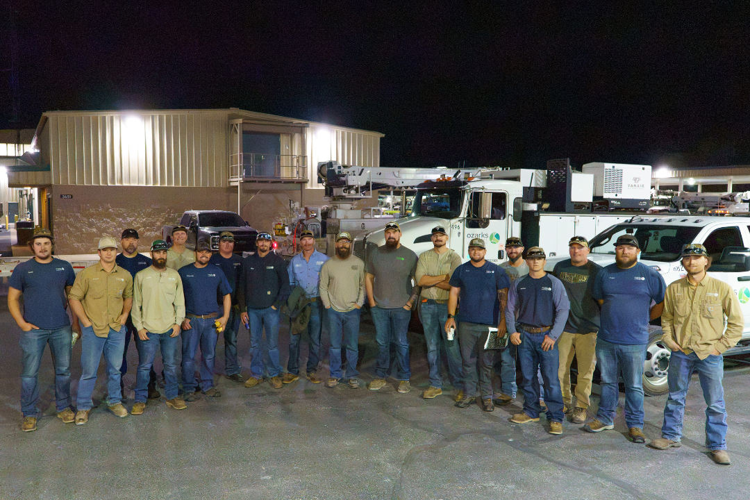 A group of workers posing outside at night, standing in front of utility trucks and a building, wearing casual work attire and hats.