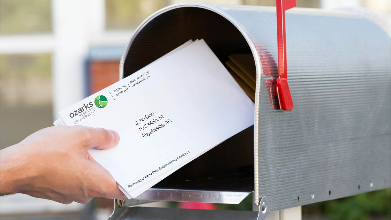 Hand placing a white envelope labeled "Ozarks" into a silver mailbox with a red flag, outdoors.