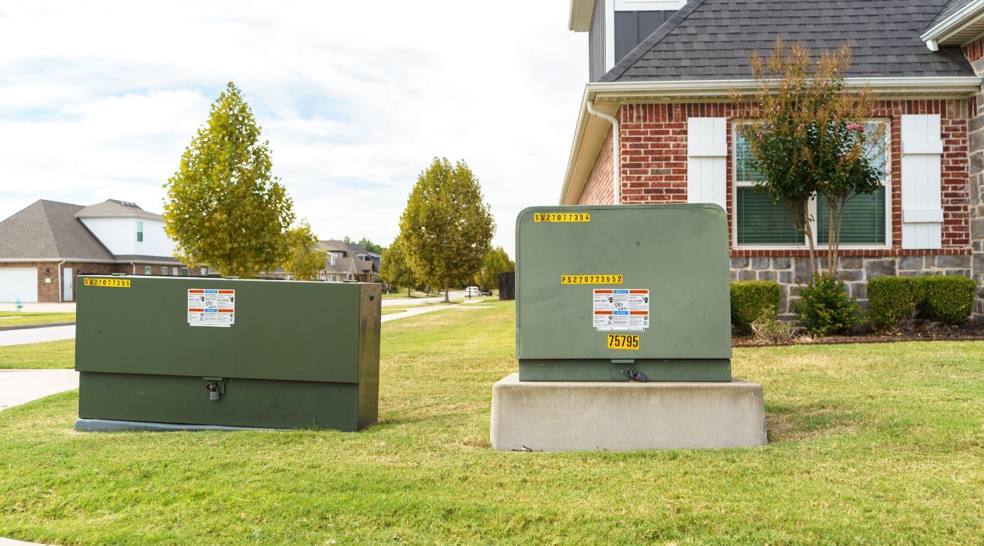 Two green utility boxes with warning labels sit on a grassy lawn outside a brick house in a suburban neighborhood.