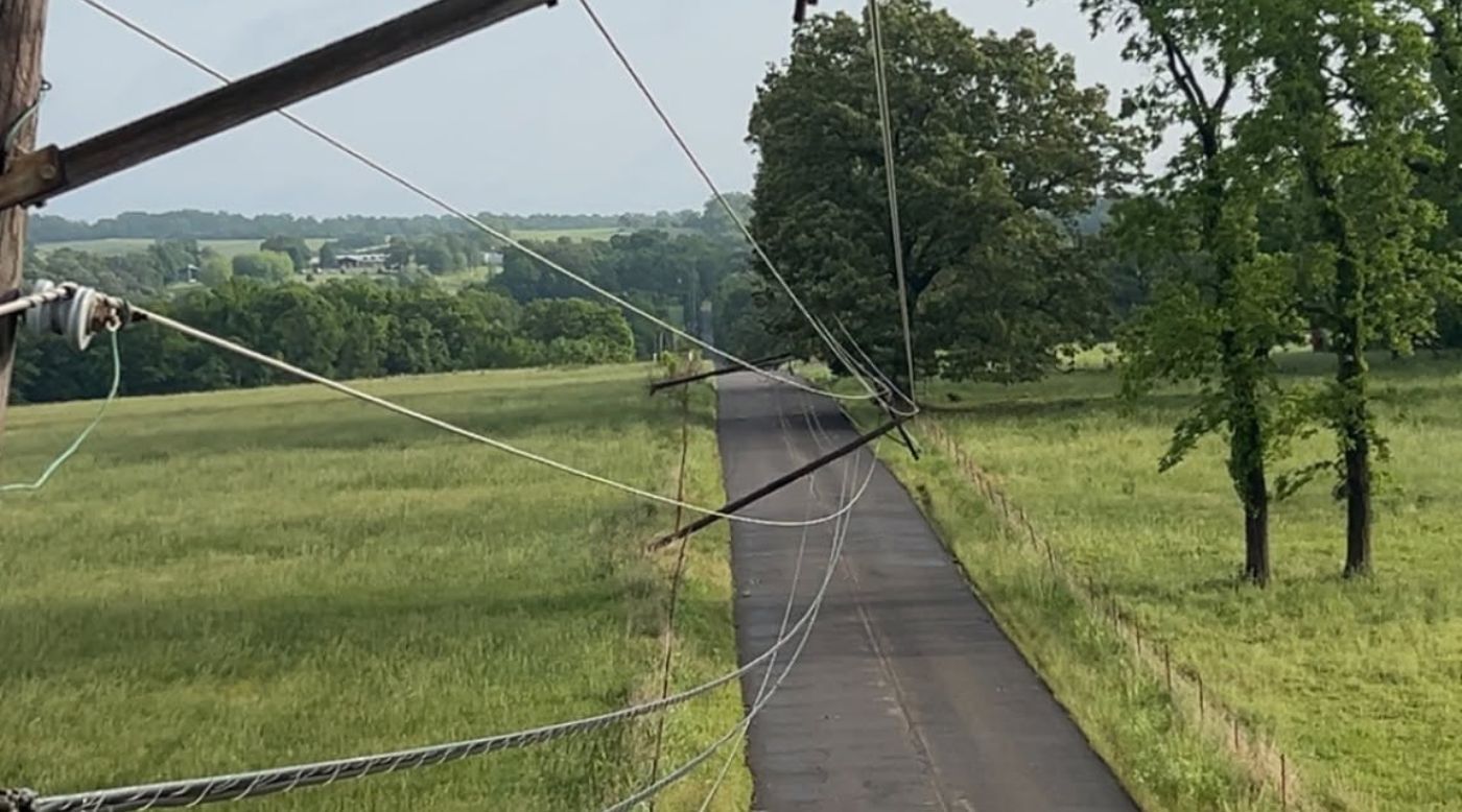 A rural road with leaning utility poles and wires, surrounded by green fields and trees under a cloudy sky.