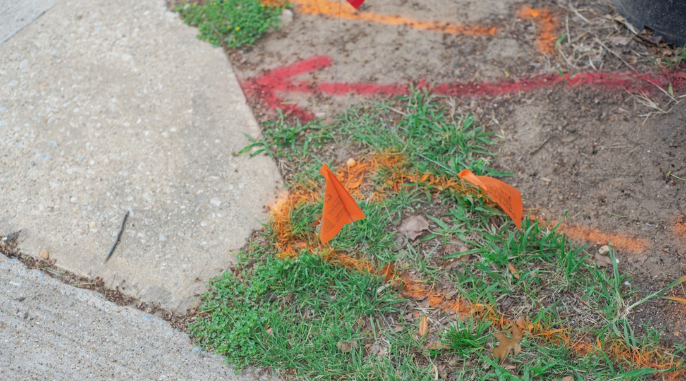 Orange and red markers on grass and dirt near a concrete path, likely indicating underground utilities or construction areas.