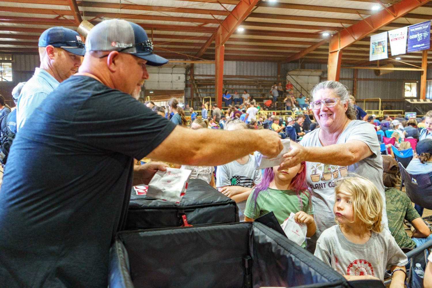 A man hands food to a smiling woman at a crowded indoor event. Children stand nearby, and people are seated in the background.