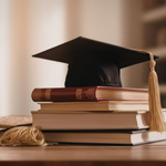 A stack of books topped with a black graduation cap and a tassel on a wooden table, symbolizing education and achievement.