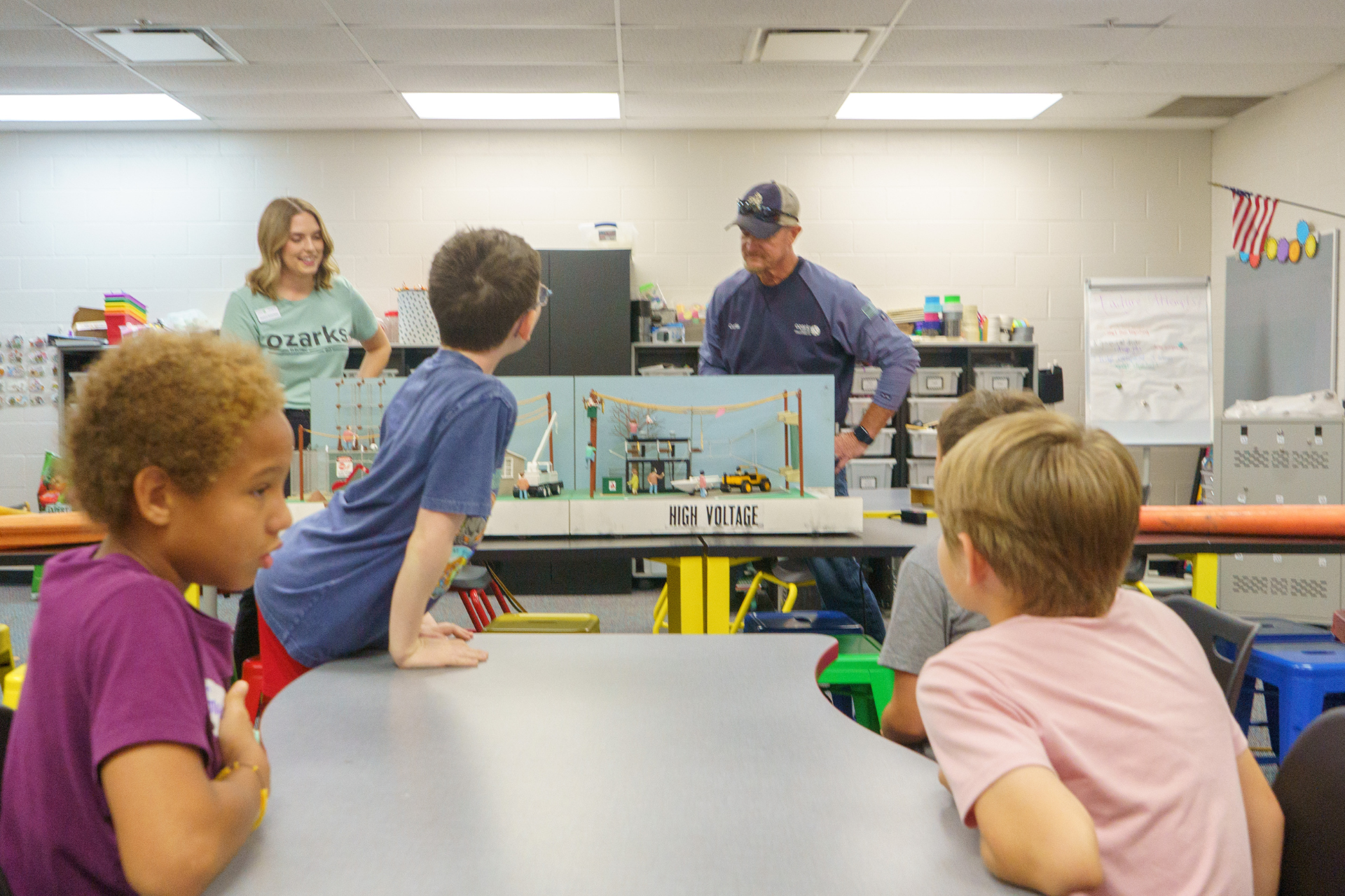 Children gather around a table in a classroom, listening to a man and a woman next to a model bridge.