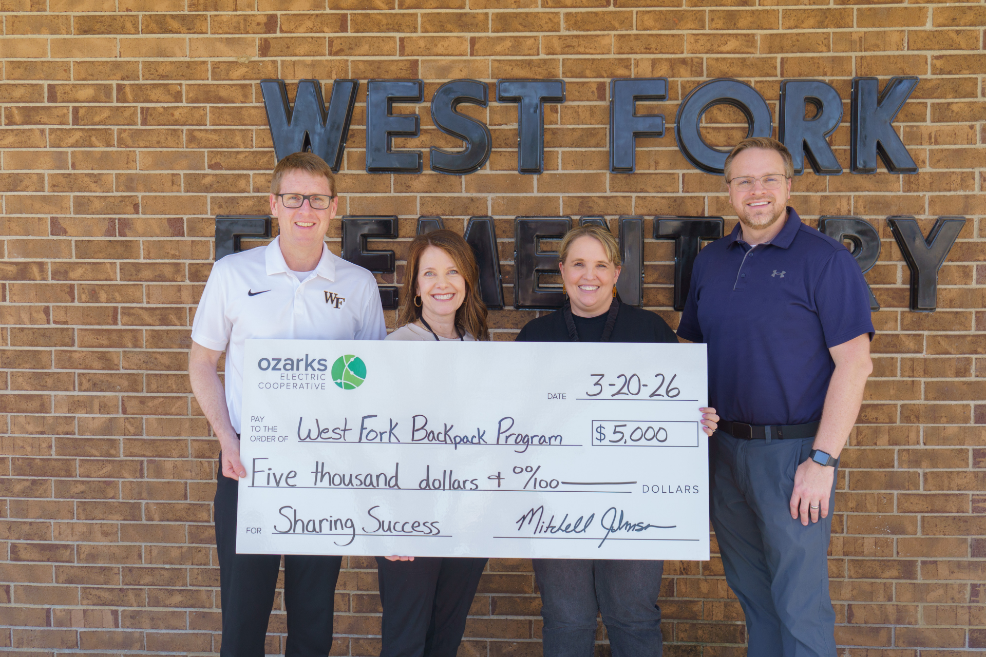 Four people stand holding a large check for $5,000 to the West Fork Backpack Program in front of West Fork Elementary.