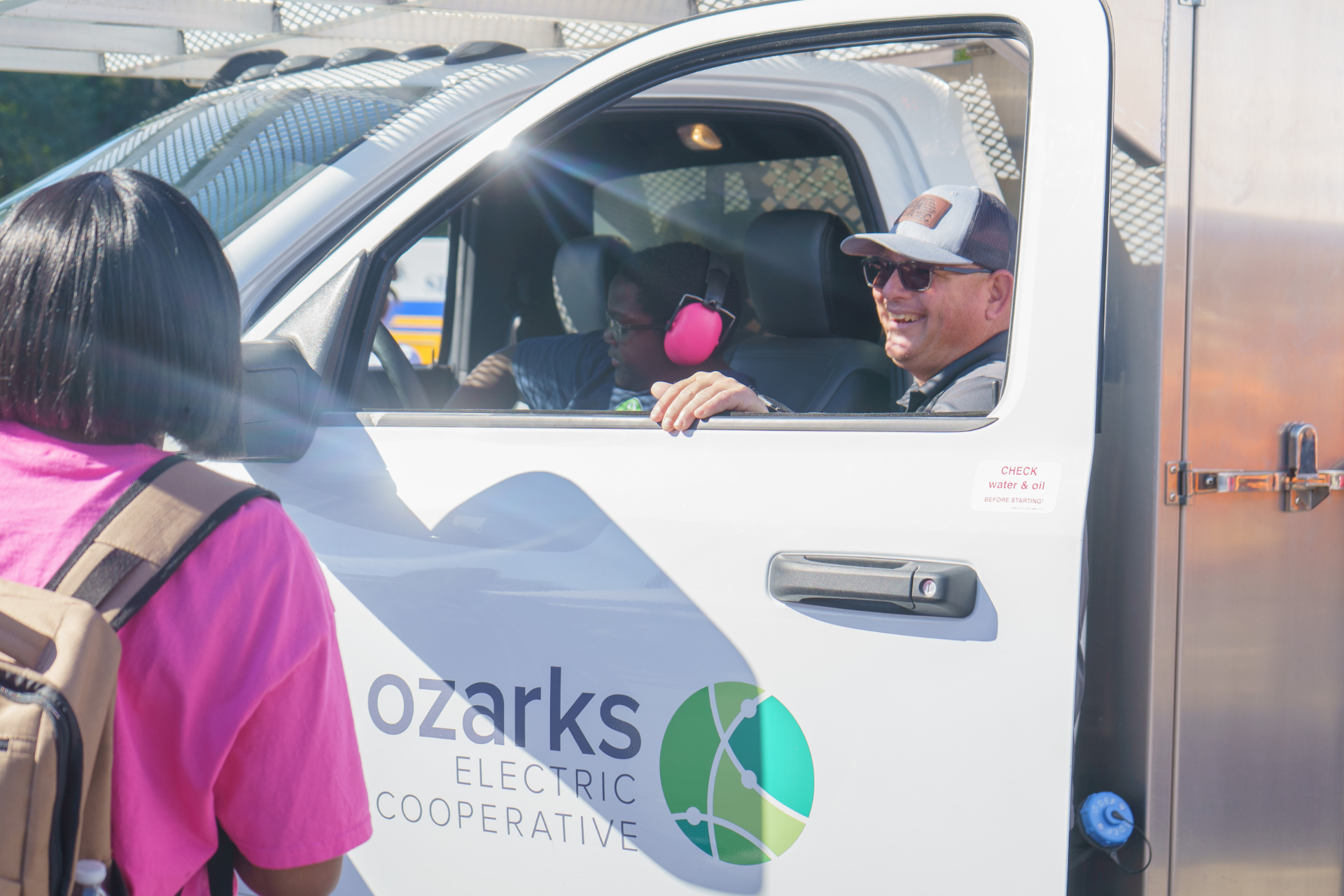 A man in sunglasses sits in an Ozarks Electric Cooperative truck, smiling at a woman with a backpack. Another person sits in the passenger seat.